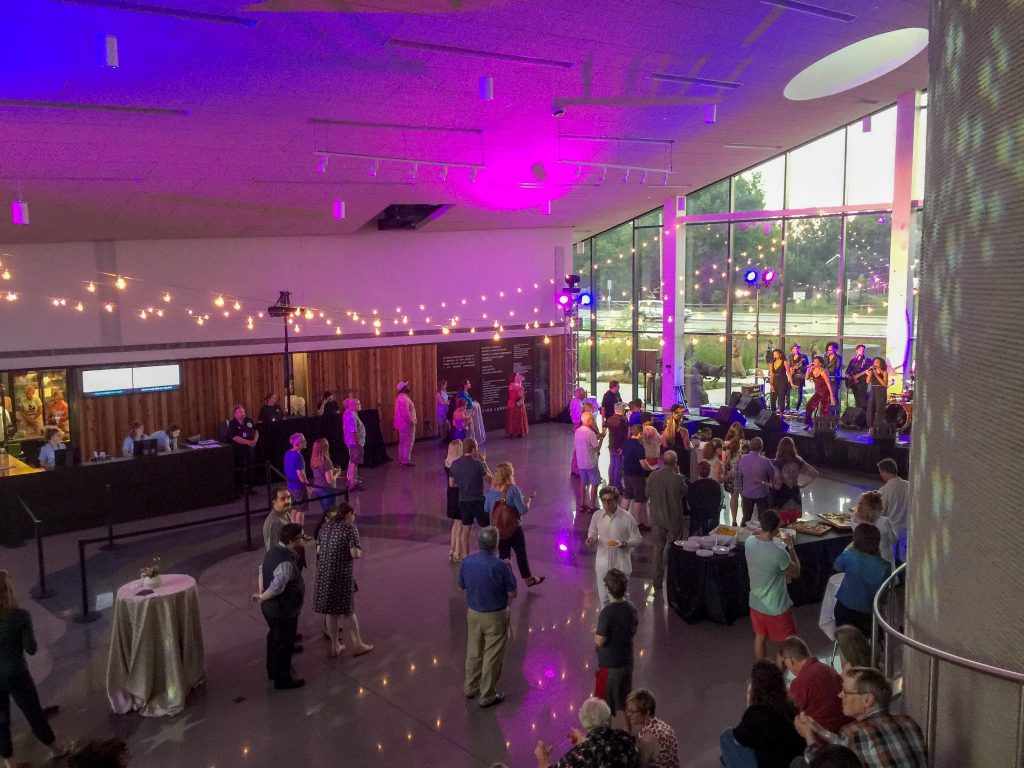 Large reception hall with standing tables and cafe lights, small crowd of people, and purple hues shining on the ceiling