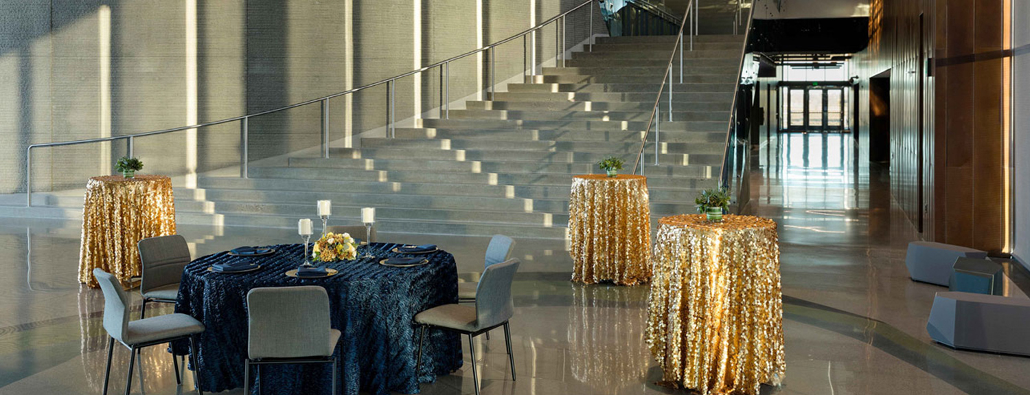 High top tables covered with sparkly gold tablecloths, round table with floral centerpiece, candles and blue table cloth, set in front of Horizon Hall's grand staircase