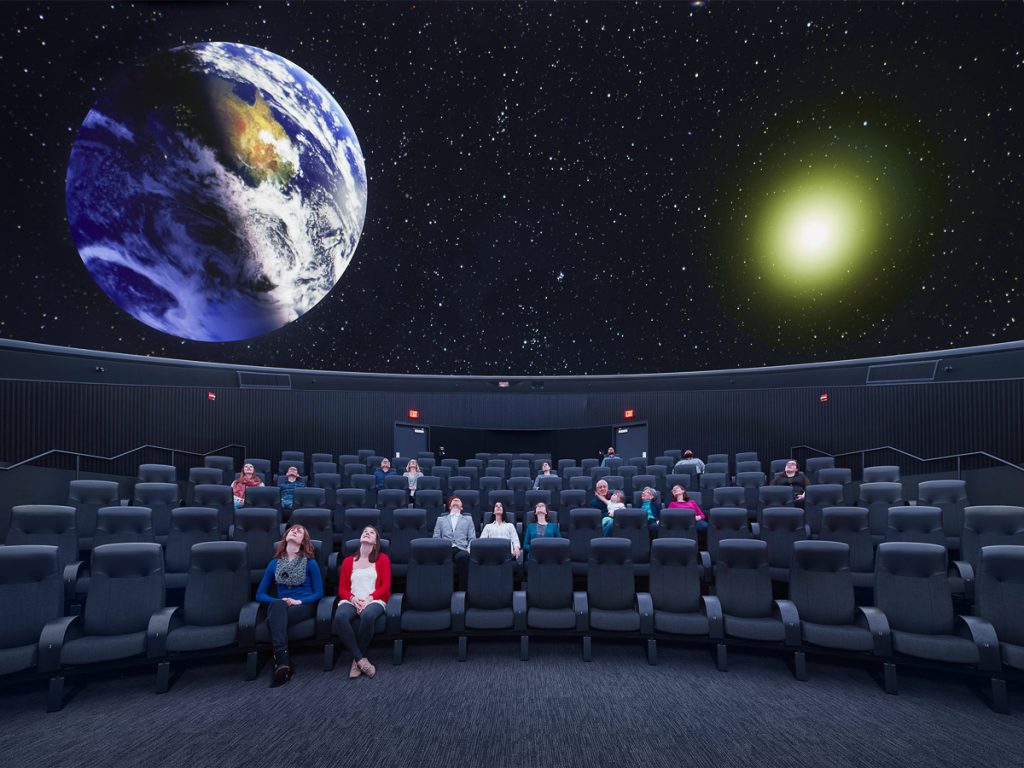 An audience looks at an image of the earth and stars projected on the planetarium screen
