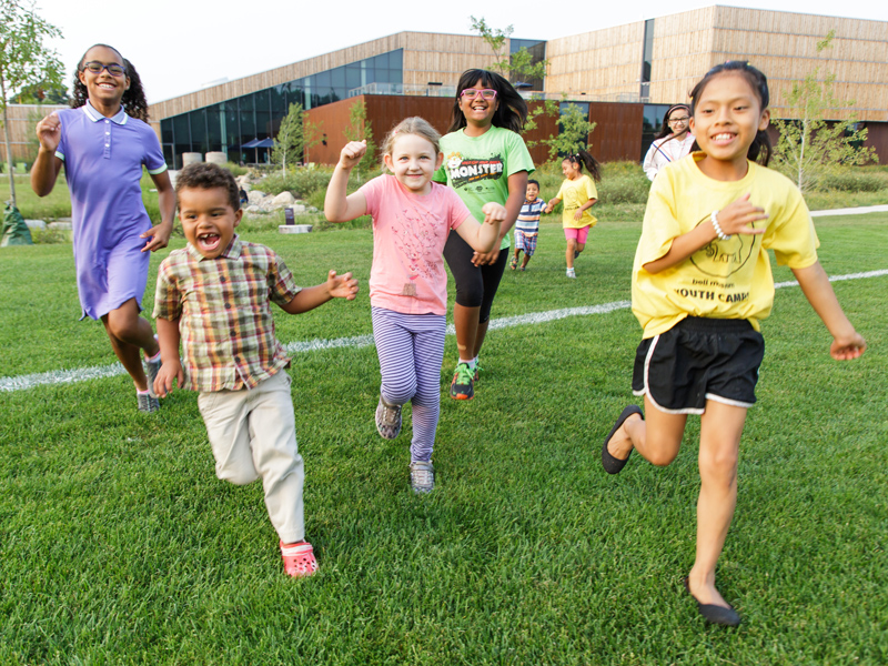 A group of kids run on the field outside the Bell Museum in the summertime