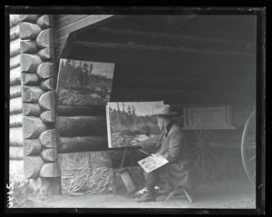 A man paints near a rustic log cabin