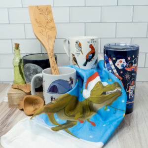 A variety of mugs, wooden kitchen utensils, and a tea towel with a t-rex in a santa hat