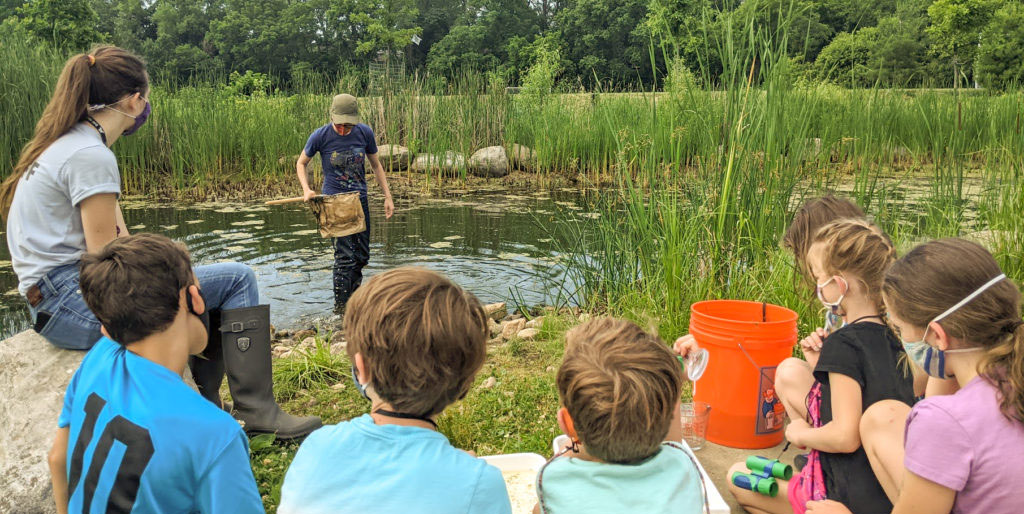 Group of children sitting on ground outside by pond listening to a presentation